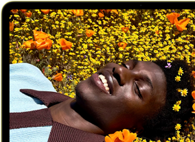 MacBook Neo, citrus colour, screen shows a smiling person lying in a sunny field of flowers